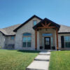 Modern brick home with a gabled roof, wooden porch beams, and manicured lawn under a clear blue sky.