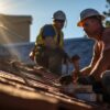 Two roofing contractors working on a roof at sunset, wearing safety gear and using tools for installation.